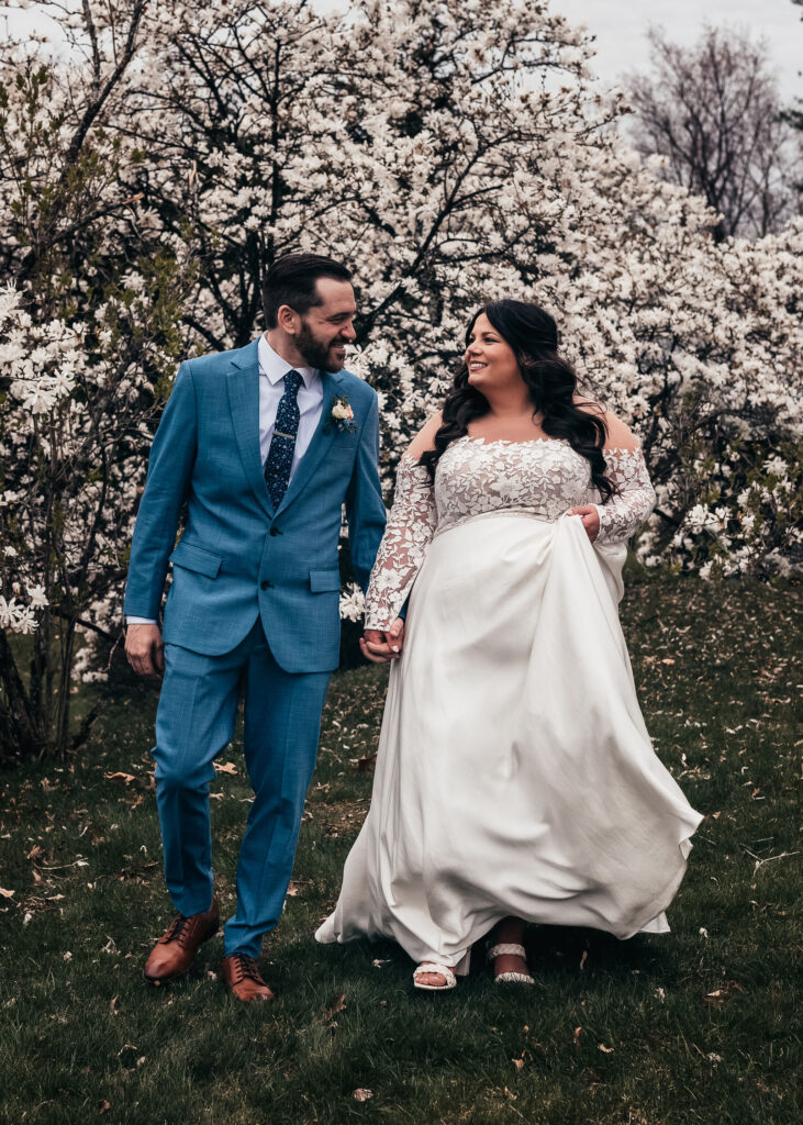 Bride and Groom walking, holding hands and having an intimate moment in front of white blossoming tree in spring wedding at Bedford Village Inn by by NH Wedding Photographer Lisa Smith Photographer
