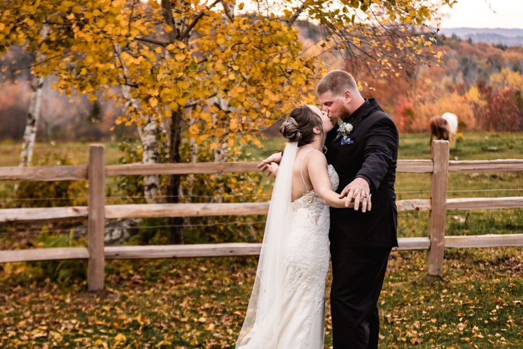 Fall wedding at Cold Spring Farm in Alton NH with bride kissing groom and horses in the background by NH Wedding Photographer Lisa Smith Photographer