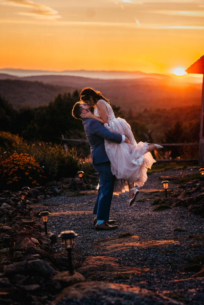 Groom lifting bride for a kiss on path in front of sunset and mountain view at Cobb Hill Estate in Harrisville NH By Lisa Smith Photography