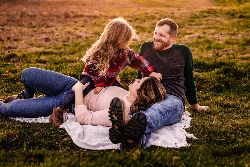 Daughter sitting on mom and tickling dads tummy in NH family photography session at Wagon Hill in Durham NH