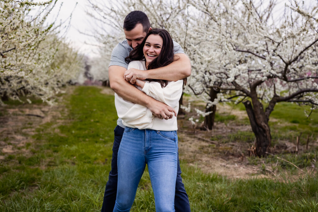 couple embracing and laughing in row of blooming apple trees at spring New Hampshire cloudy photo session at Alyson Orchard by Keene NH