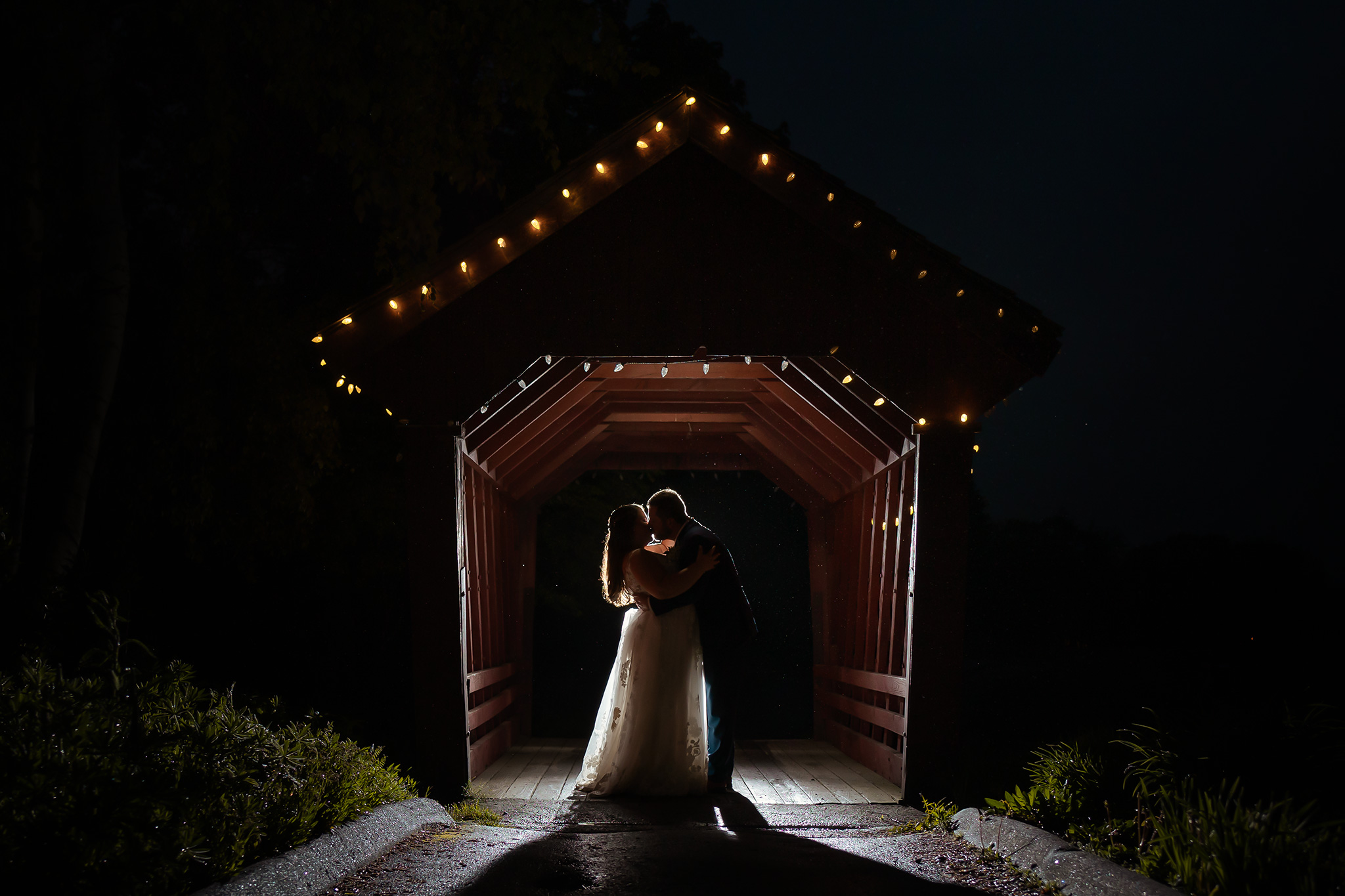 Couple kissing while being backlit and framed by covered bridge in NH Wedding Photography creative night photography at The Farmhouse at Candia Woods. 