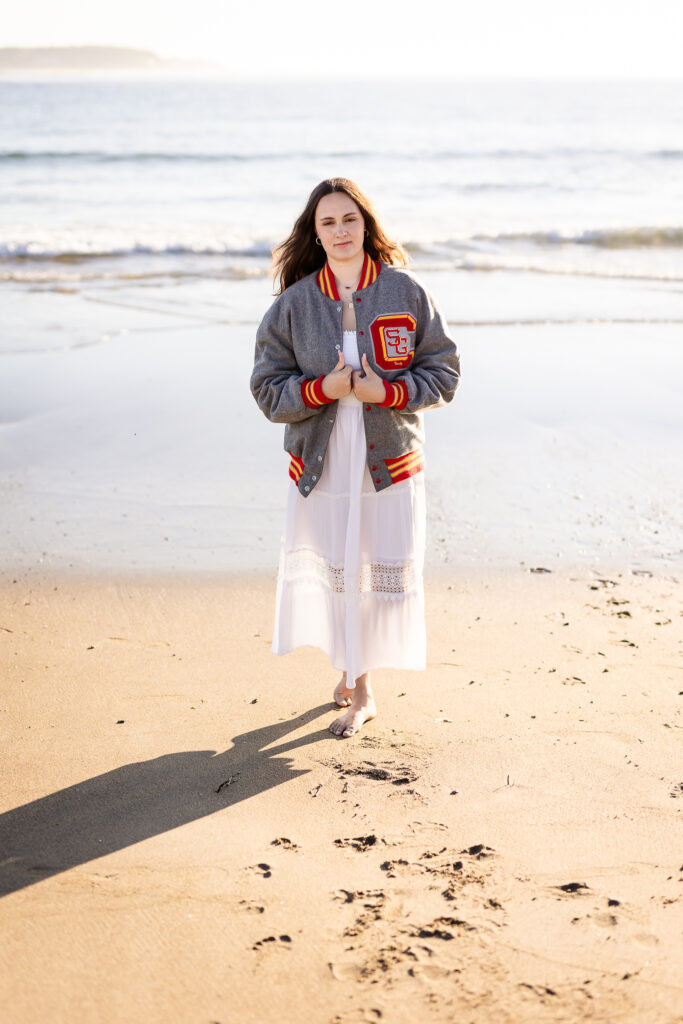 High school senior girl walking on beach with her senior varsity jacket at Shorts Sands in York Maine by NH Senior Photographer Lisa Smith Photography