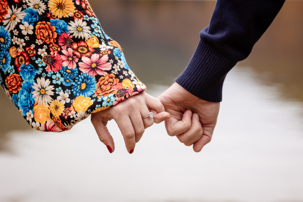 Couple holding pinky fingers to show engagement ring, women is wearing floral pattern dress and guy is in a navy blue sweater by NH engagement photographer Lisa Smith Photography