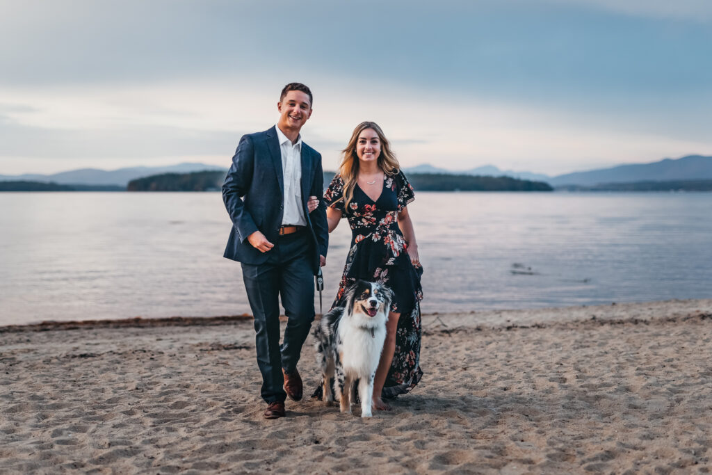 Couple walking dog on beach with mountain views, wome wearing beautiful navy blue floral print dress and guy is in navy blue suit by NH engagement photographer Lisa Smith Photography