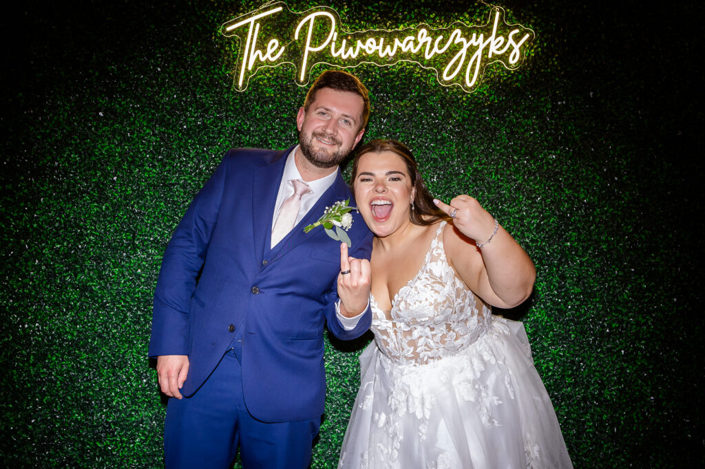 Bride and groom showing off their rings in front of green ivy wall with a neon sign of their name.