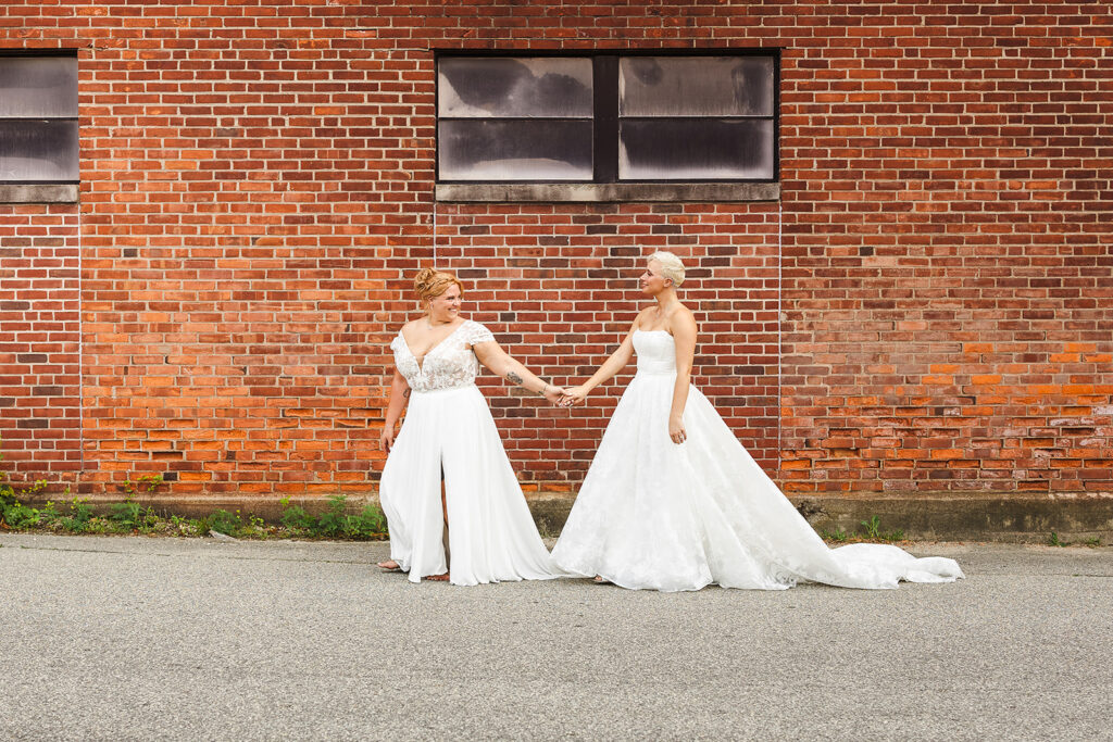 Two brides, both in beautiful white wedding gowns walking hand in hand in front of a red brick wall.