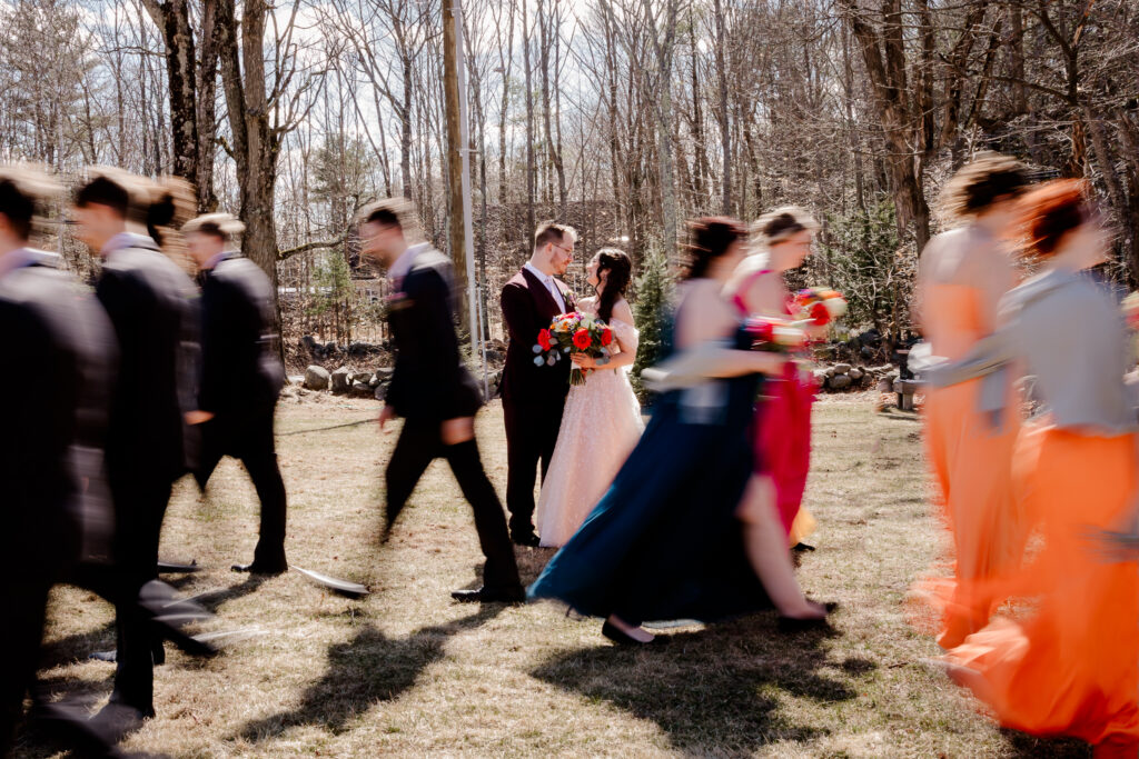 Bride and groom looking at each other while bridal party walks past them in a blur at the Barn on Merry Hill wedding in Nottingham NH